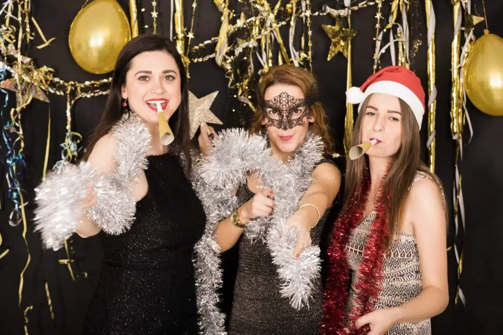 Groupe d'amies s'amusant avec des accessoires de Noël et guirlandes argentées devant un photobooth à Paris.